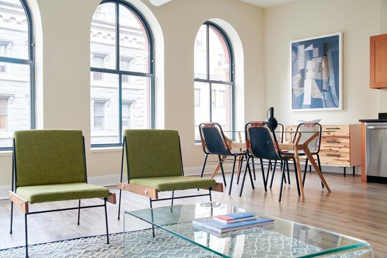 Modern loft space with green accent chairs, plexi coffee table, modern dining set, large arch windows, light wood floors.