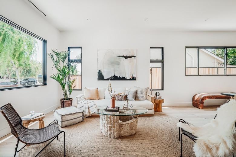 Living room with a round fiber rug, glass and stone coffee table, white sofa with multi-colored pillows, windows of varied sizes, and a palm