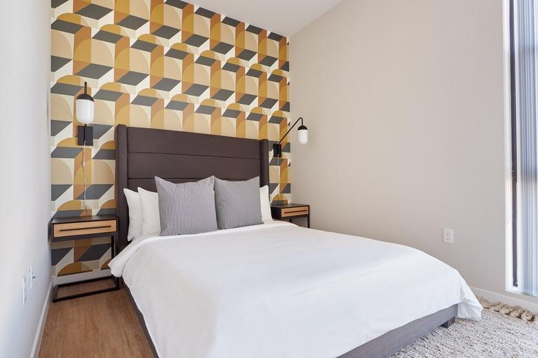 Eclectic bedroom with dark brown fabric headboard, gold and grey geometric wallpaper, metal and glass sconces, nightstands, gray pillows, rug.