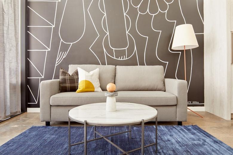 Modern loft living room with gray couch, pillows, floor lamp with white lampshade, concrete floor, round marble and metal coffee table, art mural on wall, sheer curtains.