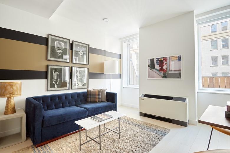 Modern living room with navy blue couch, glass coffee table, side table,  brass lamp, beige black and white striped wall, rug, large windows.