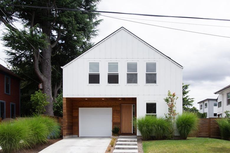 Contemporary house, with a white and brown exterior. A concrete step walkway, and plants line the grass lawn.