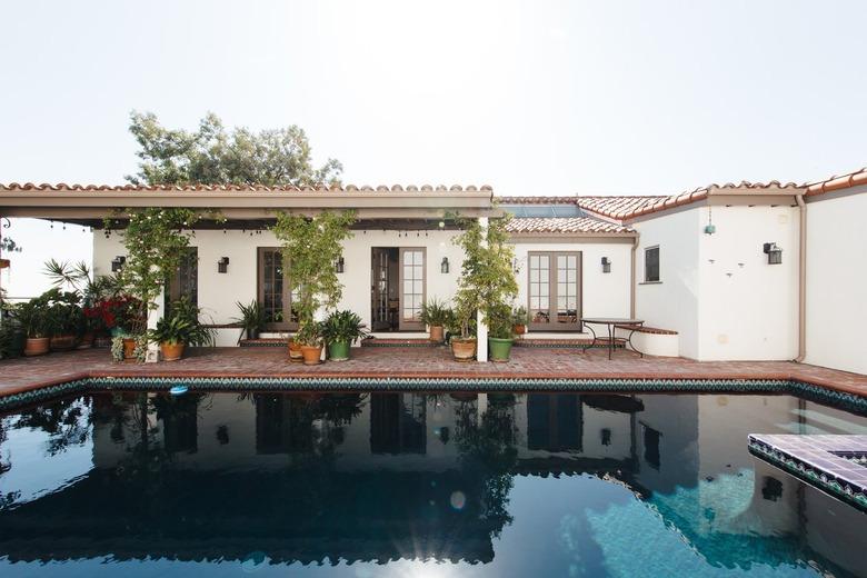 White Spanish house, with terra cotta roof tiles and skylights. Pool with blue tiles. Turquoise patio chairs, and plants in planters.