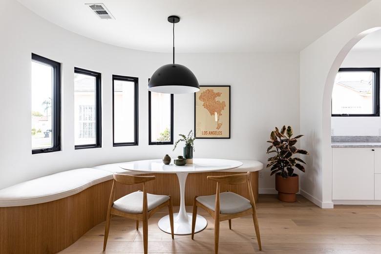 Contemporary curved dining room with curved bench, small round table, chairs, pendant lamp, framed print, and tall windows with view of hallway via arch