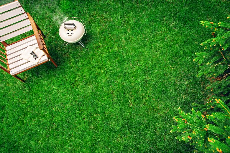 Ivory colored grill on the grass near the wooden armchair with a book and glasses. Top view