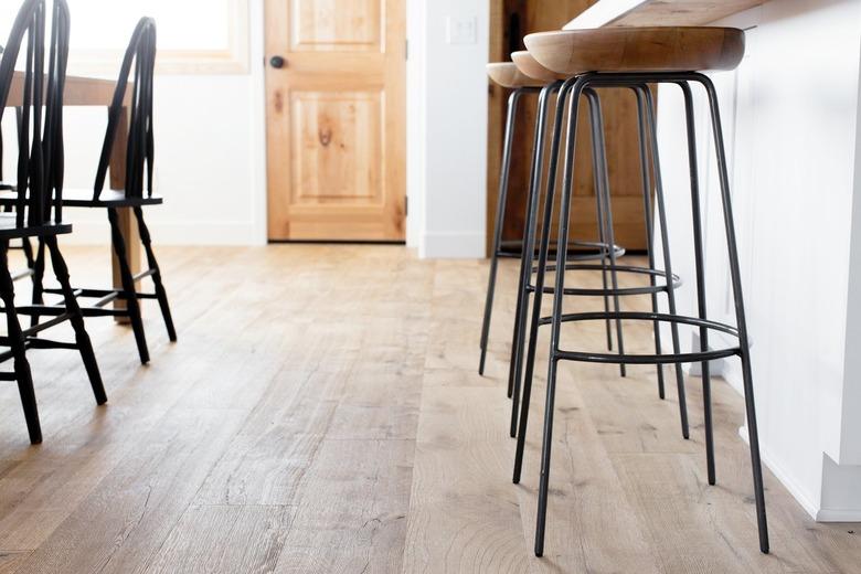 Chairs between kitchen and dining area with a pathway between leading to a wooden door. The floor is wood and the walls are white. On the right, three bar stools with wooden seats and metal legs. On the left, black Windsor chairs.