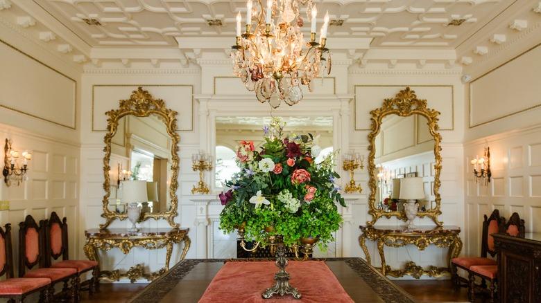 A very formal dining room with tons of architectural details. A large crystal chandelier hangs over an ornate dining table with a tall flower arrangement in the center. There are formal chairs lined against the wall and two ornate gold console tables with mirrors flank the fireplace.