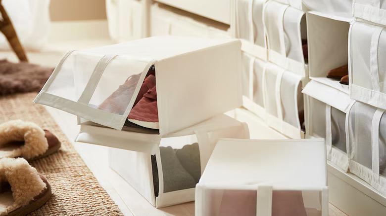 three white IKEA SKUBB shoe boxes on the floor, one open revealing a pair of shoes, with many other SKUBB boxes stacked up on the closet shelves behind them