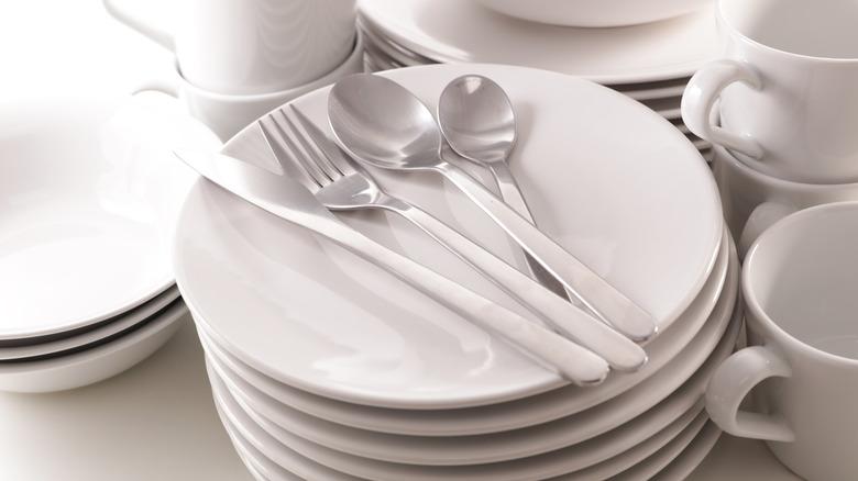 stack of white ceramic dinner plates with a setting of four pieces of silverware on top and surrounded by stacks of bowls, other plates, and mugs