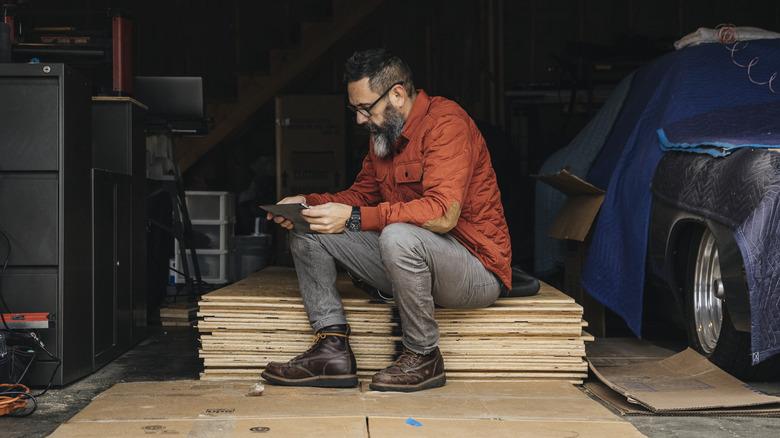 Man consulting tablet while sitting on stack of plywood