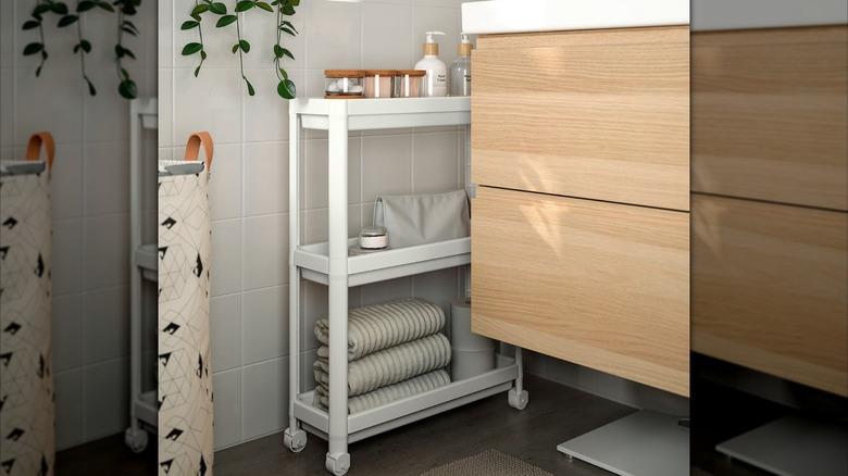 white IKEA VASKEN cart with extra bathroom towels, products, and toilet paper rolled part way out in a narrow space between the wood bathroom vanity and white tiled wall