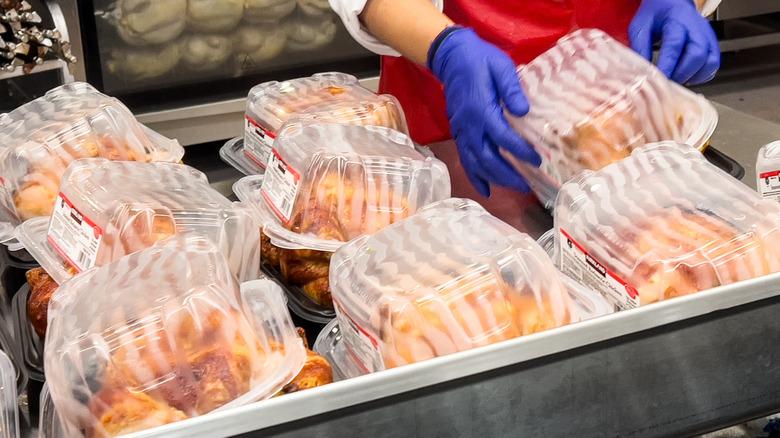 gloved hands holding rotisserie chickens in plastic-topped containers at a grocery store