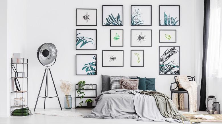 A white bedroom with high ceilings. There is a gallery wall behind the bed. Each picture is framed with black and has plants or insects on a white background. The room has a bed, chair, shelves, and a tripod lamp.