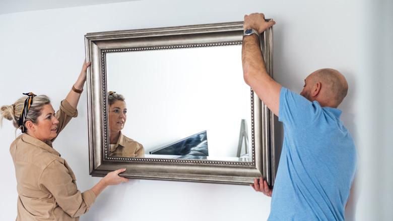 A middle-aged man and woman are hanging a large mirror with a silver frame on a wall.