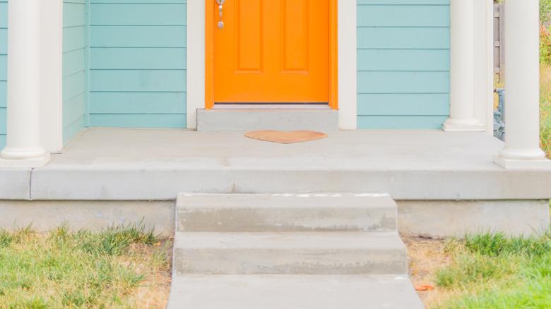 Close-up in the entrance to a home with pale blue-green painted siding, white trim, and an orange door.