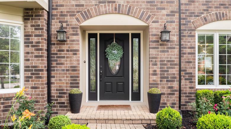 The entryway of a brown brick home with off-white trim and a black front door. The yard is beautifully landscaped and there are black planters on either side and a spring-time wreath on the door.