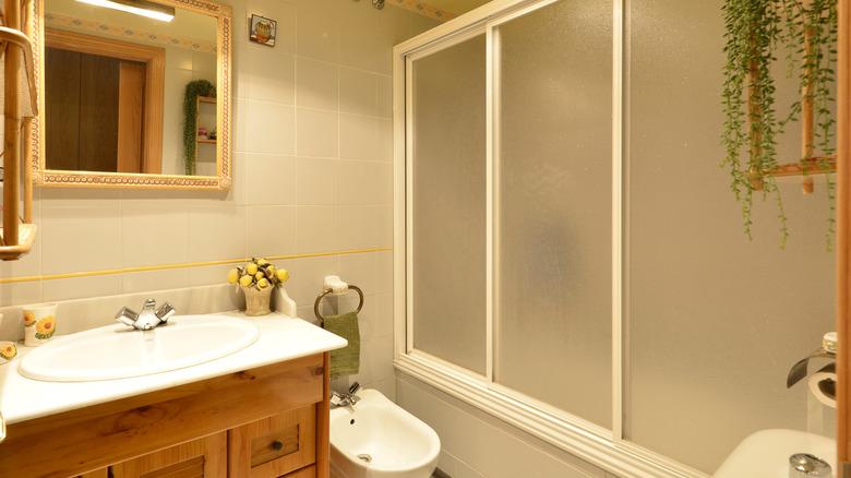 An older bathroom with wood features includes a framed glass shower door with frosted glass.