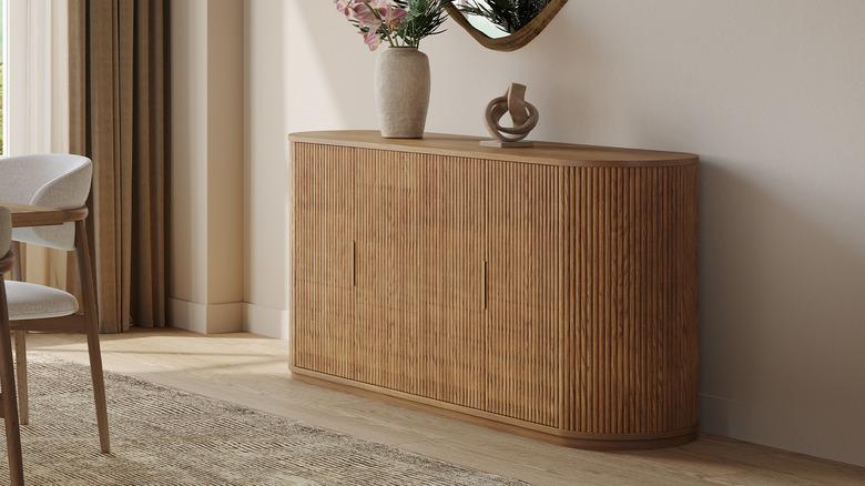 Dining room with off-white walls, light wood floors, cream area rug, taupe drapery, and a natural wood sideboard with a reeded texture and curved sides