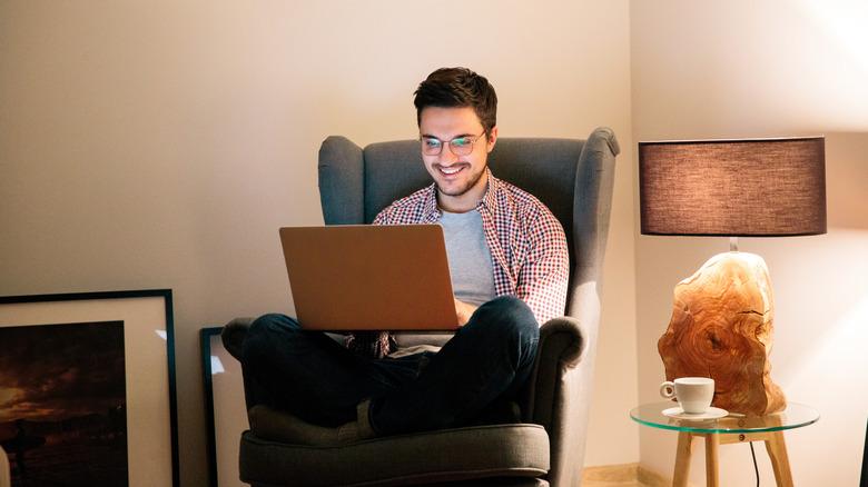 Young man sitting on a chair in his living room, looking at his laptop with a big smile on his face. The room is illuminated by the warm glow of a nearby lamp, and a cup of coffee sits on the end table.