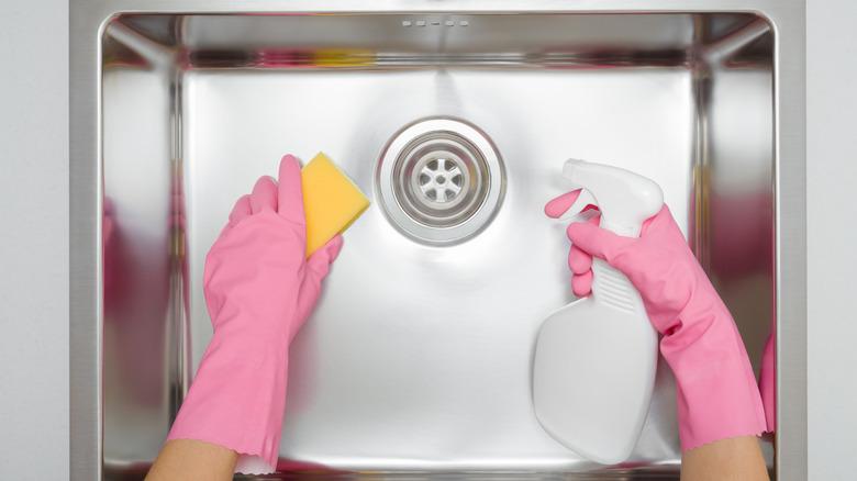 shot of someone wearing pink gloves holding a sponge and a spray bottle and cleaning a stainless steel sink