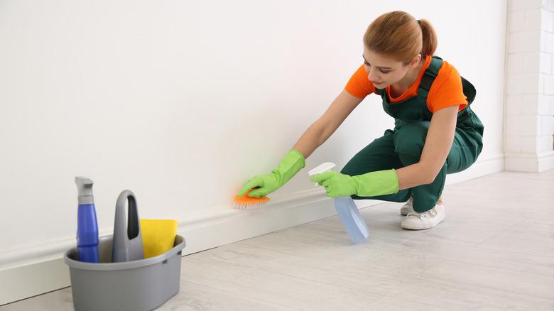 A young woman in green overalls is bending at the knees to clean a baseboard. She is wearing cleaning gloves and holding a scrubbing brish and spray bottle. A caddy filled with cleaning supplies is nearby.