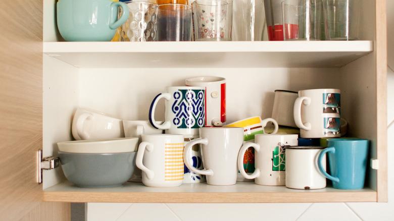 interior of open upper cabinet with an unorganized assortment of mugs stacked and piled on the lowest shelf