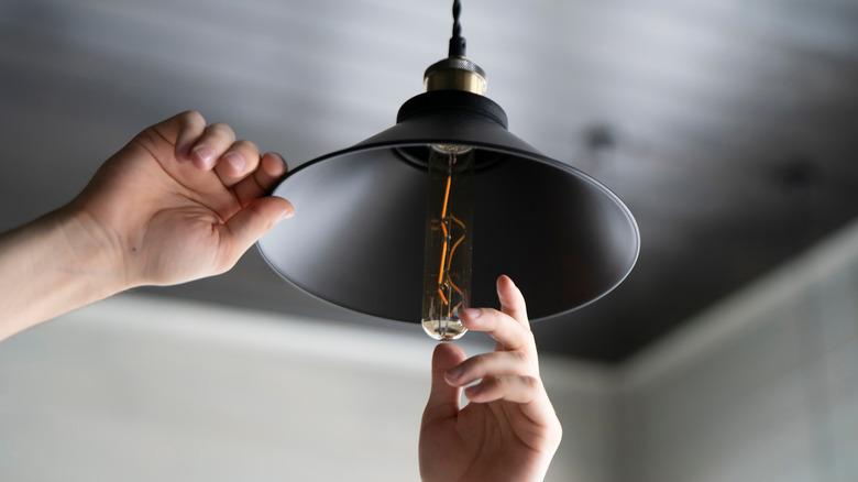 man changing a tubular-shaped exposed filament light bulb in black light fixture with a black shade