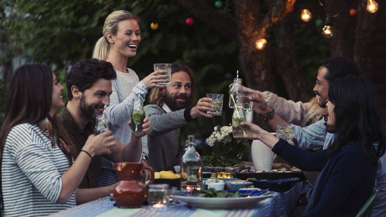 group of young adult friends sitting at an outdoor table with outdoor lighting, all holding mocktails and toasting each other