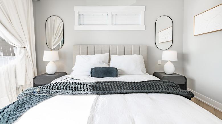 A neutral bedroom is painted light gray with white trim, white curtains, white bedding, and white lamps. Nightstands, throw pillow and throw blanket are dark gray, and headboard is light gray.