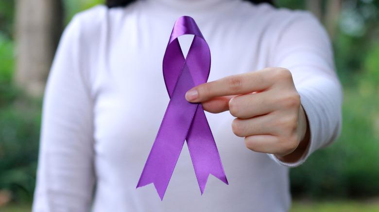 Someone in a white shirt holds out a purple domestic violence awareness ribbon for the camera.