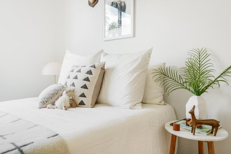 White bedding, black-white patterned pillows, stuffed animal and blanket. Stool with geometric vase of palm leaves and a sculpture