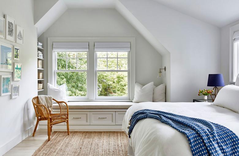 attic apartment with window seat, cane chair, white walls, white bedding, blue throw.