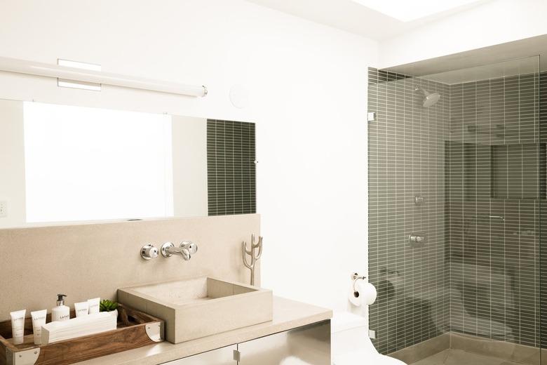 Bathroom with a square sink basin and facet fixed to a wall. A fluorescent bar light above. Shower with glass door and gray wall tiles.