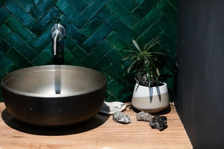 Wood counter with black bowl sink next to potted plant against green tile wall