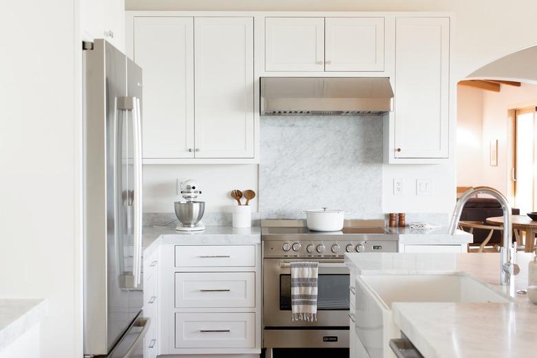 Kitchen with a stainless steel range, white cabinets, and a marble countertop. On the countertop, there's a white KitchenAid stand mixer with a stainless steel mixing bowl along with a white utensil holder with wooden salad tongs. On the stovetop, there's a white dutch oven. To the left, there's a stainless steel refrigerator. To the right, there's a white kitchen island with a sink with a chrome faucet.