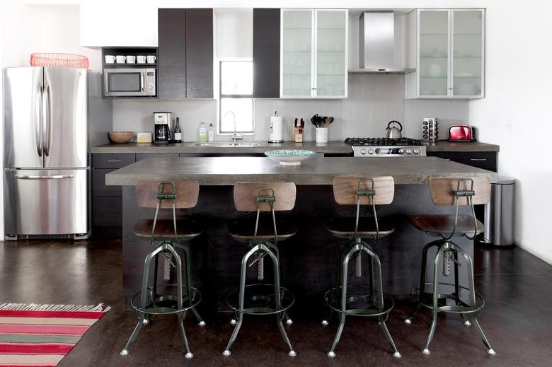 A kitchen with frosted cabinet doors, dark wood cabinets, gray kitchen island, industrial bar stools, and a colorful striped rug.