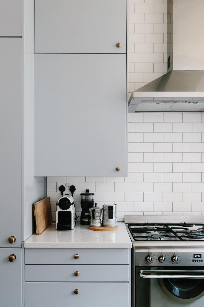 White tiled backsplash with grey kitchen cabinets, white countertops, silver stovetop, and various kitchen supplies