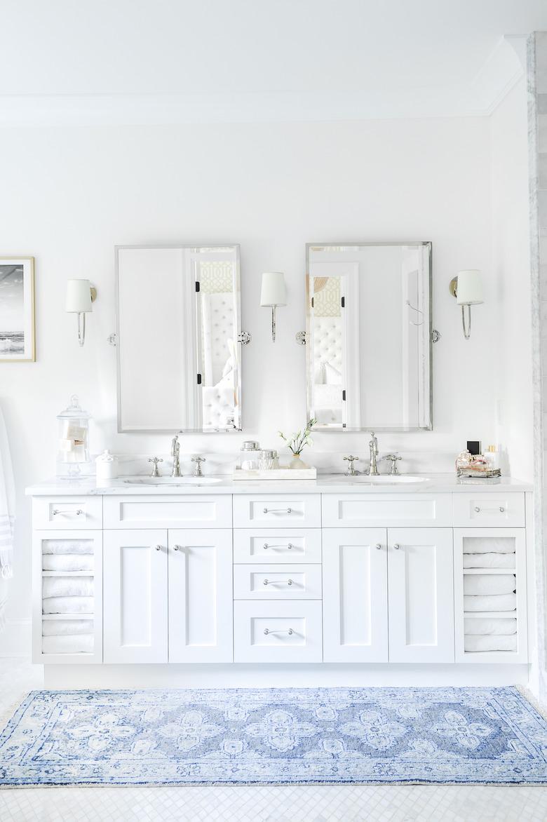 bathroom with white cabinets, marble backsplash, two mirrors, and blue rug