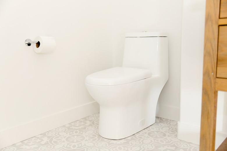 A white toilet in a bathroom with white walls and a white and grey tiled floor. A roll of toilet paper is on the wall next to the toilet, hanging on a simple chrome holder.