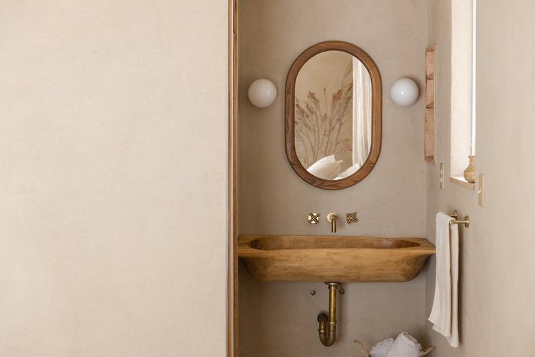 Minimalist bathroom with a wood oval mirror, globe sconces, wood sink basin, gold faucet hardware, and towel bar.
