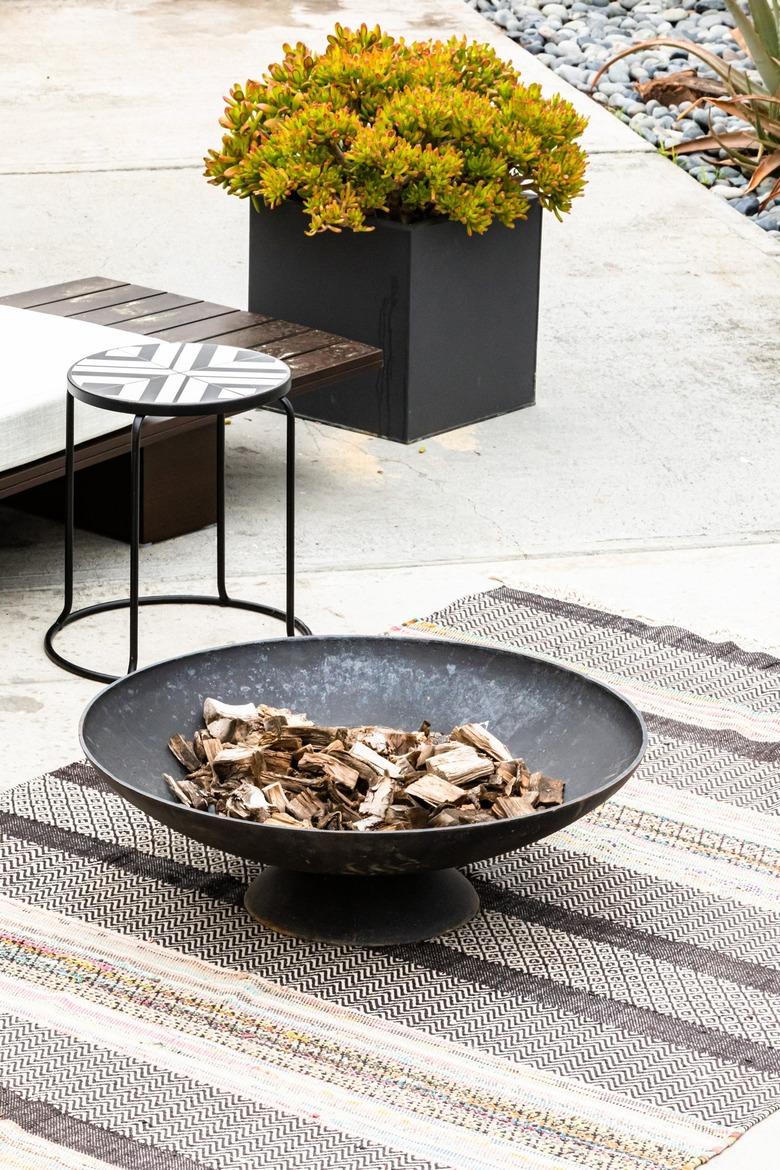 A patio fire pit on a gray-neutral striped rug with a plant and stool