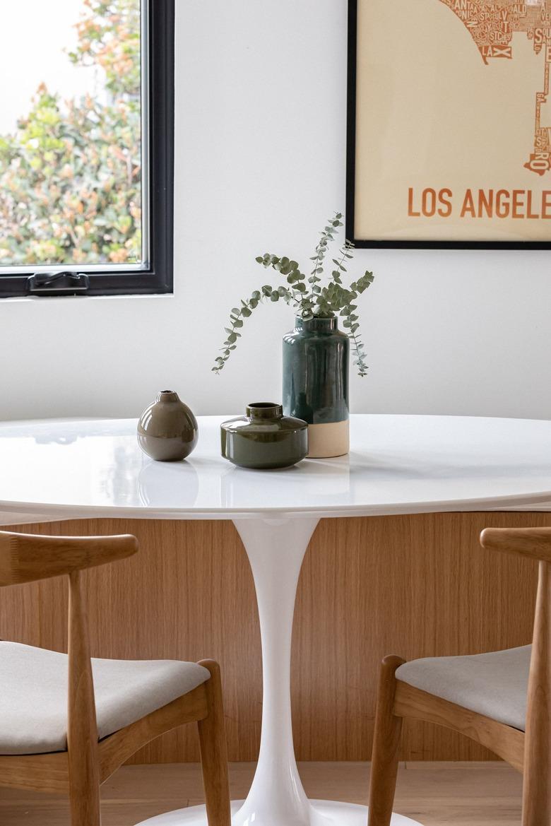 Modern dining room with white round table with plant, chairs, curved wood bench, and framed print against white wall with window