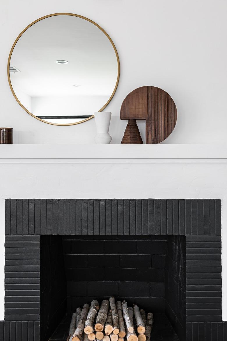 White mantle above black fireplace with wood sculptures next to round mirror against white wall