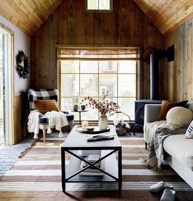 Cabin living room with couch, plaid chair, coffee table, pillows, throws, wood panel walls.