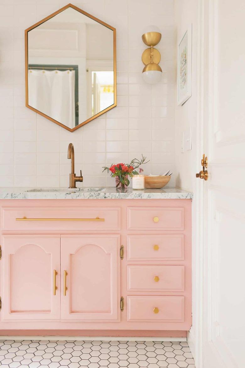 French country bathroom sink in bathroom with pink vanity, marble sink and counter, brass faucets, mirror and light.