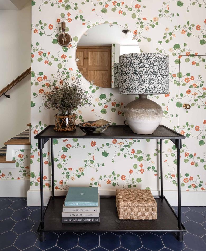 Entryway with wallpaper, lamp, black metal table, books, basket, plant, round mirror.