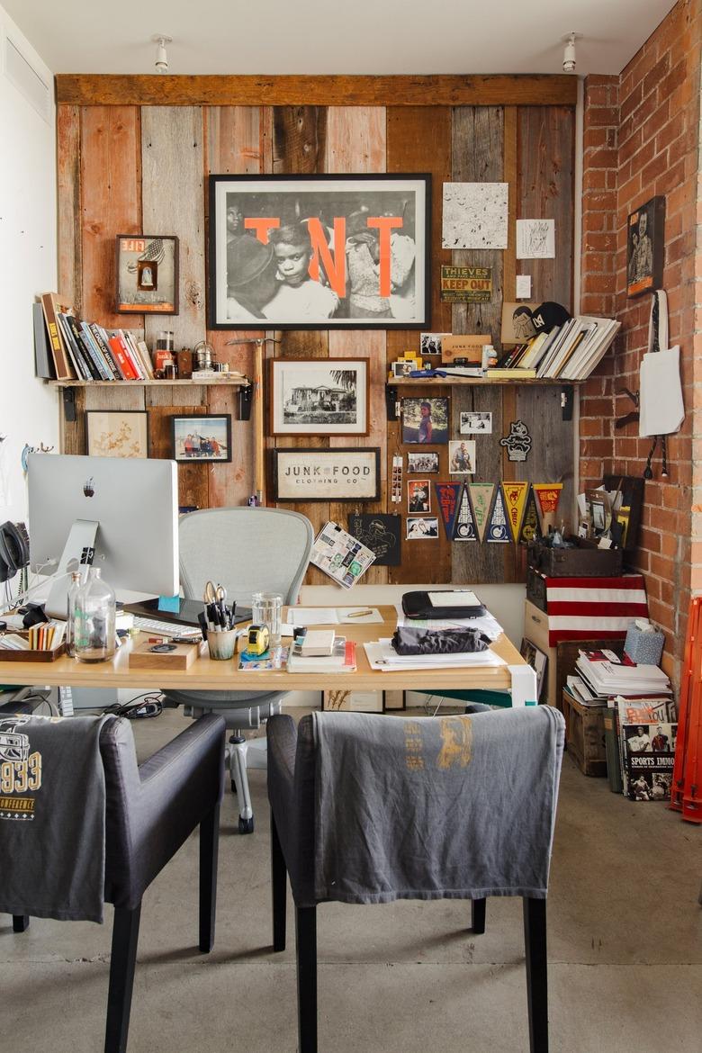 An office with varied wood panel and brick walls, eclectic art, shelves, wood desk, computer, and black chairs.