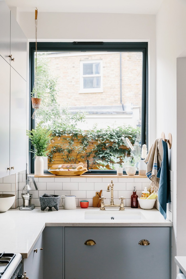 White kitchen with white countertops, grey cabinets, and large window revealing back patio with wood fencing and plants