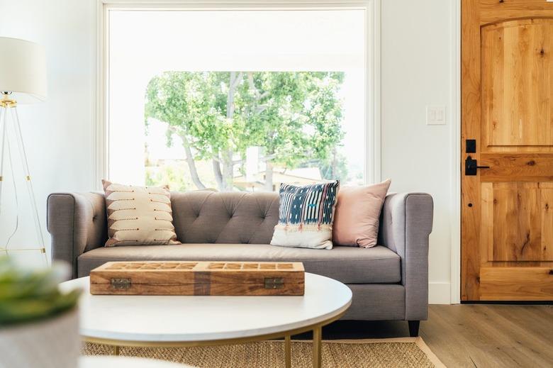 A gray tufted upholstery sofa with multi-colored pillows by a picture window and a wood door. A round white coffee table and wood floor.