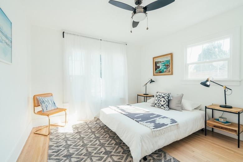 Californis-bohemian style bedroom with black and white rug, cane side chair, white curtains in window, side tables with black lamps, and a ceiling fan.